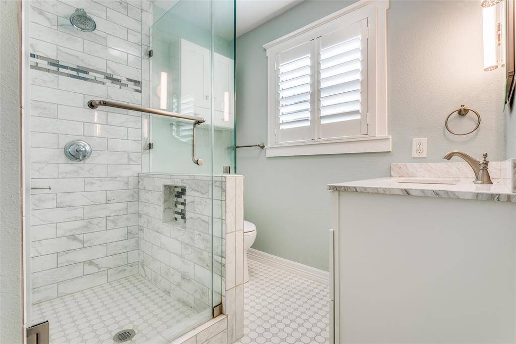 This is a modern bathroom featuring a walk-in shower with beautiful tile, a glass door, a white vanity, and a window with white plantation shutters. The space is accented with brushed metal fixtures and a soothing green wall color.