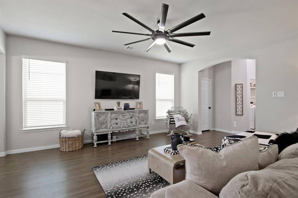 The spacious living room features a modern ceiling fan, large windows for natural light, neutral-toned walls, and wood look vinyl flooring, creating a cozy and inviting atmosphere. An open archway leads to the front door & garage entrance, enhancing the flow of the space.