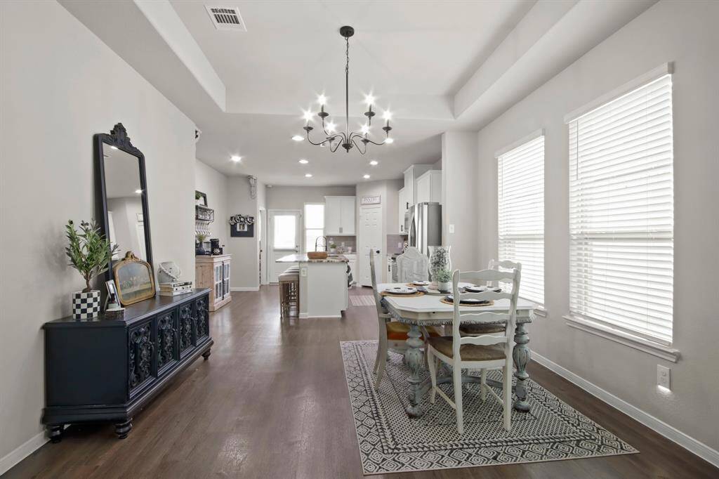 This photo showcases a bright, open-concept dining and kitchen area. The space features light walls, dark wood look vinyl flooring, and a stylish chandelier. The large windows allows natural light to flood the space. In the background, a modern kitchen with white cabinetry and stainless steel appliances is visible.