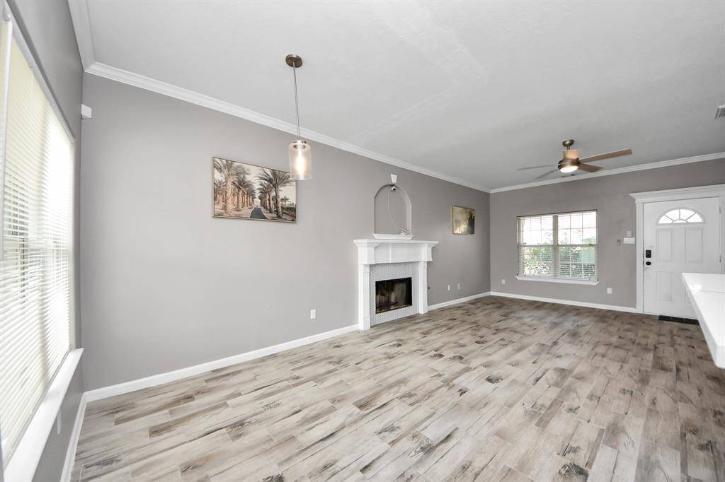Bright living room has modern flooring, neutral gray walls, and a cozy fireplace. Features a ceiling fan, large windows for natural light.