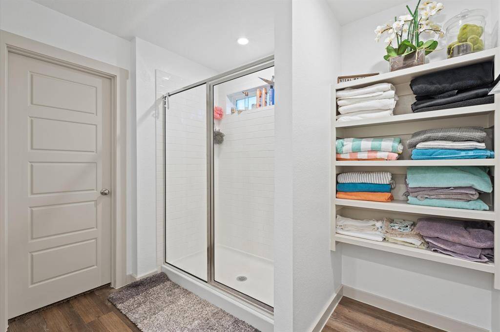This is a modern, bright bathroom featuring dual sinks set in a spacious white vanity with ample storage. There's a large mirror above the vanity and an open shelving unit for towels and bathroom essentials. The room also includes a walk-in shower with a glass door and a separate toilet area behind a closed door. The flooring is a wood-look laminate, providing a nice contrast to the white walls and fixtures. Approximate Measurements:11X8
