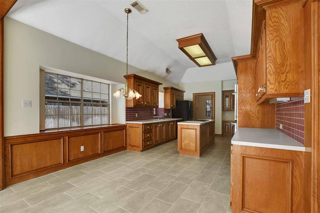 This kitchen features ample natural light, wooden cabinetry, a central island, and tiled flooring. A large window offers a view outside.