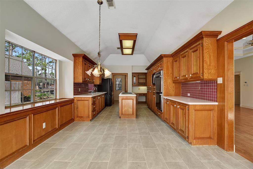 This kitchen features a spacious layout with abundant wooden cabinetry, modern appliances, and a central island. Large windows offer plenty of natural light, while the decorative light fixture adds charm. The tile flooring is both stylish and practical, complementing the overall warm aesthetic.