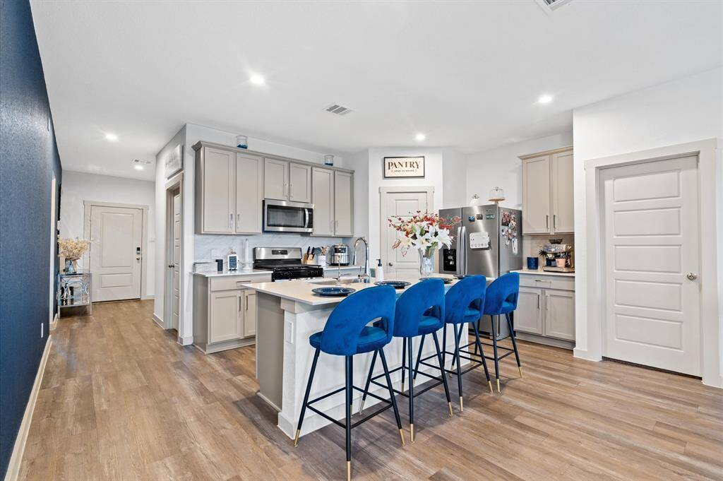 This is a modern, spacious kitchen featuring stainless steel appliances, light gray cabinetry, and a central island with plenty of room for barstools. The room has premium laminate flooring and includes a designated pantry area. Approximate Measurements: 17x11