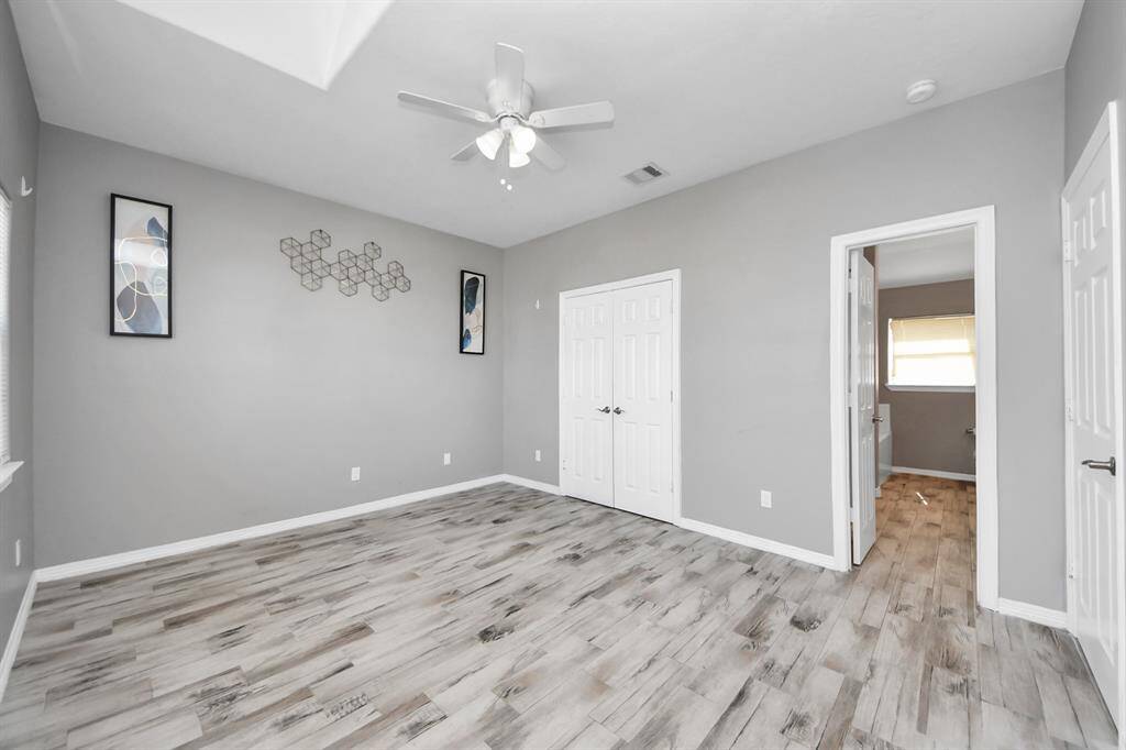 The primary bedroom features modern gray walls, stylish wood-look flooring, and a ceiling fan. It includes a closet with double doors and an adjoining bathroom, providing convenience and comfort. Natural light brightens the space, enhancing its welcoming atmosphere.