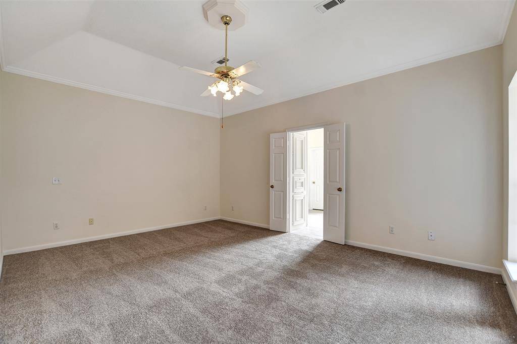 This spacious primary bedroom features neutral walls, plush carpeting, a ceiling fan, and double doors leading to the ensuite bathroom, offering ample natural light and a cozy ambiance.