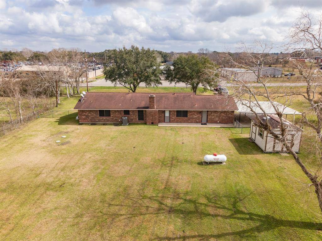 This photo shows a single-story, brick house with a large, open backyard. It features a brown roof and a small outbuilding inside the fenced in area. The area is spacious and open.