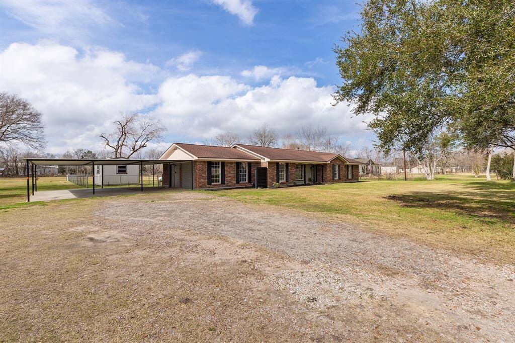 This photo shows a single-story brick home with a large, open front yard. The property features a carport and is surrounded by mature trees, offering a spacious and serene setting.