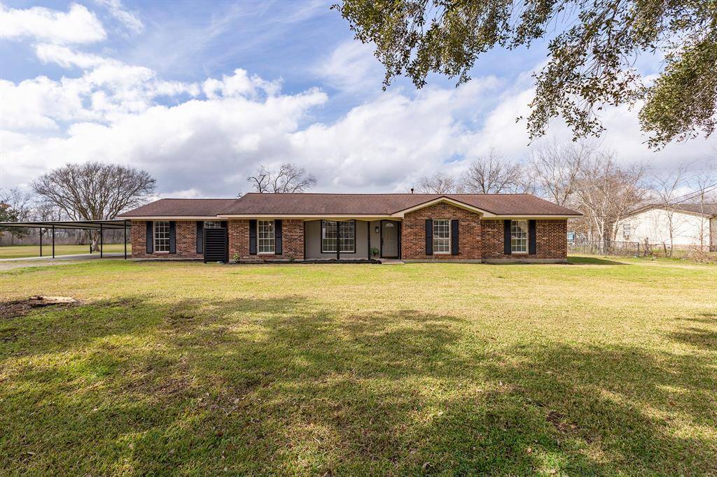 This photo features a charming single-story brick home with a spacious front yard. It has a classic design with multiple windows, a covered entryway, and a carport on the side. The property is surrounded by mature trees, providing a serene and inviting atmosphere.