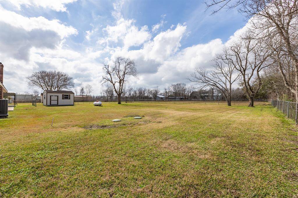 Spacious backyard featuring a wide open grassy area with a few mature trees, a small storage shed, and a propane tank. The clear sky above enhancing the sense of openness.