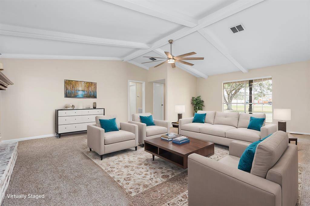 This spacious living room features a high vaulted ceiling with exposed beams and a ceiling fan. It's bright and inviting, with large windows and neutral-toned walls. The room is virtually staged with modern furniture, including a comfortable sofa set, a coffee table, and decorative accents.
