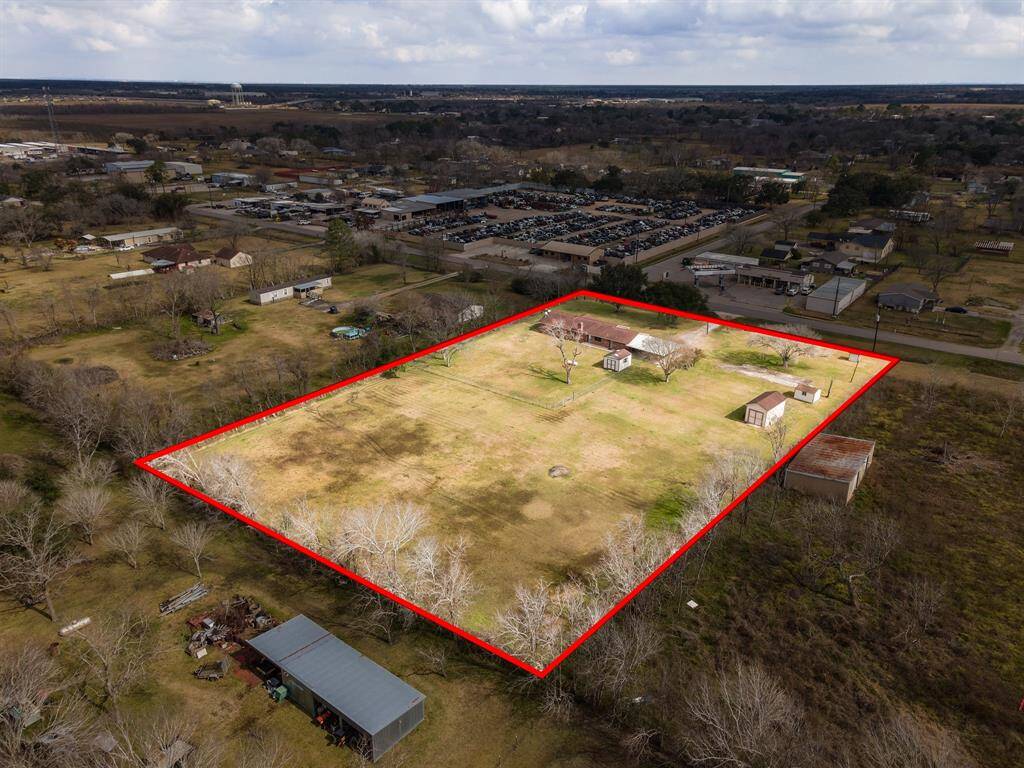 This photo shows a large rectangular property outlined in red, featuring a spacious open field with a few scattered trees and several small structures, including a house. It's situated in a semi-rural area with nearby buildings and roads, offering a balance of open space and accessibility.