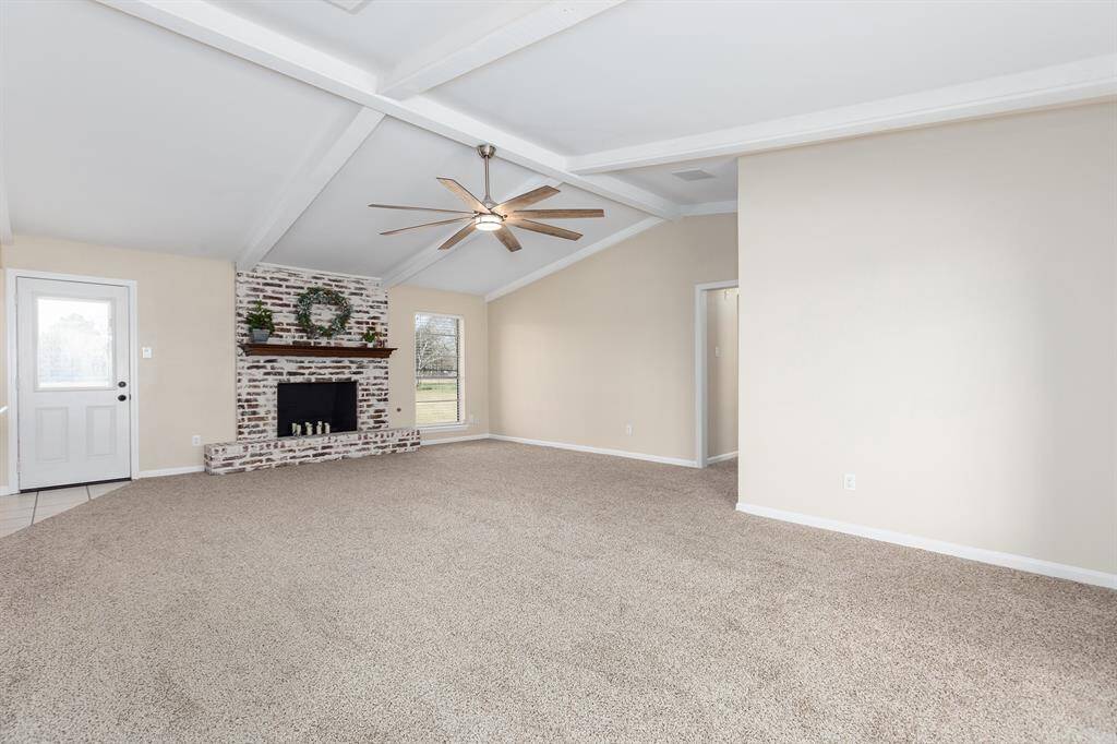 Spacious living room with vaulted ceilings, featuring a cozy brick fireplace, modern ceiling fan, and ample natural light from large windows. Neutral carpet and wall colors create a warm, inviting atmosphere.