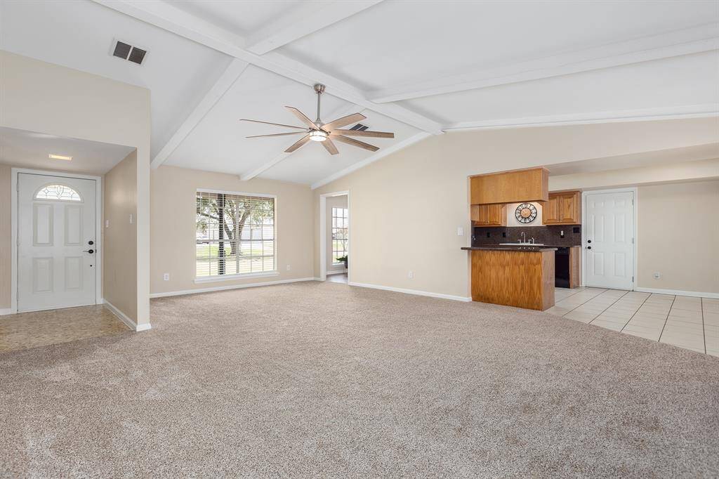 This spacious living area features vaulted ceilings with exposed beams, a stylish ceiling fan, and large windows offering plenty of natural light. The carpeted floor transitions to a tiled kitchen with warm wood cabinetry, creating an inviting open-concept space.