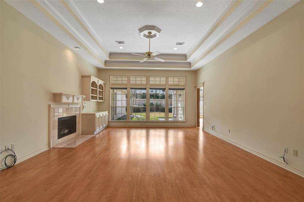 This spacious room features a hardwood floor, high tray ceilings with recessed lighting, and a ceiling fan. It has a fireplace with built-in shelving and large windows overlooking the backyard, providing abundant natural light.