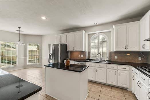 This kitchen features white cabinetry, black countertops, and stainless steel appliances. It has a central island, a bright breakfast nook with large windows, and tile flooring, creating a spacious and inviting atmosphere.