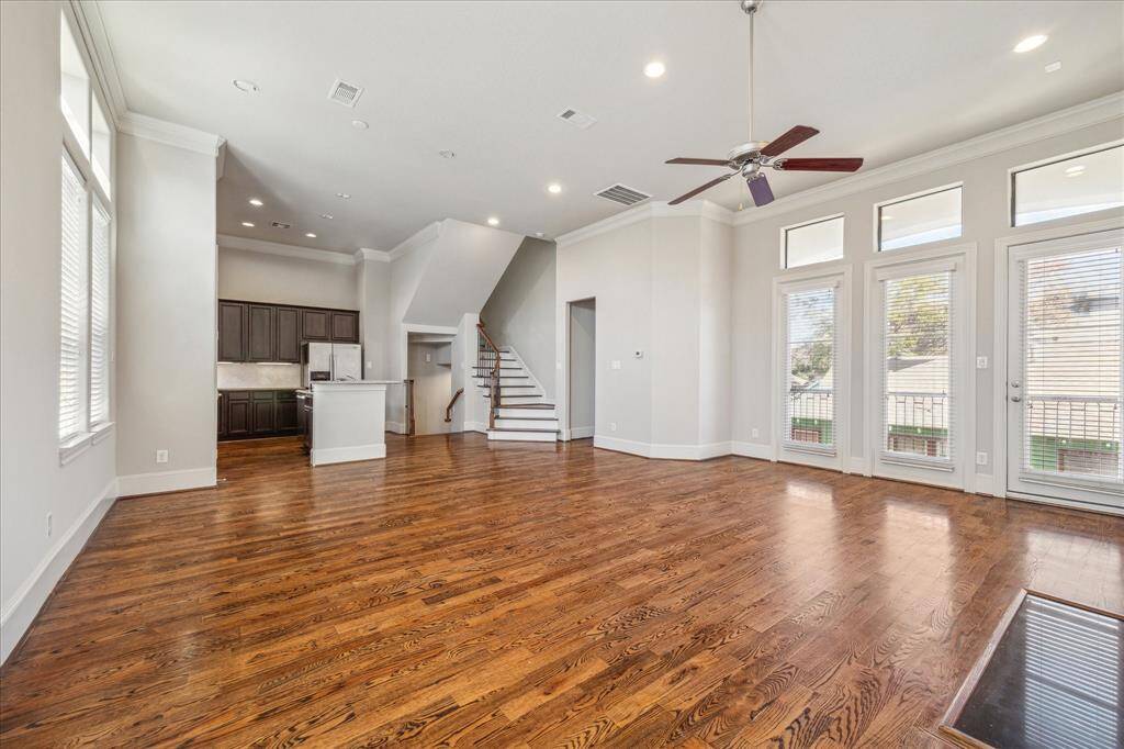 This photo showcases a spacious, open-concept living area with beautiful hardwood floors, high ceilings, and ample natural light. It features a modern kitchen with dark cabinetry, a staircase leading to the upper level, and large windows with patio doors opening to a balcony.