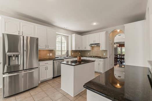 This kitchen features modern stainless steel appliances, white cabinetry with pull out shelving and dark countertops. It includes a central island, tiled flooring, and an arched entryway leading to a dining area, creating an open and inviting space.