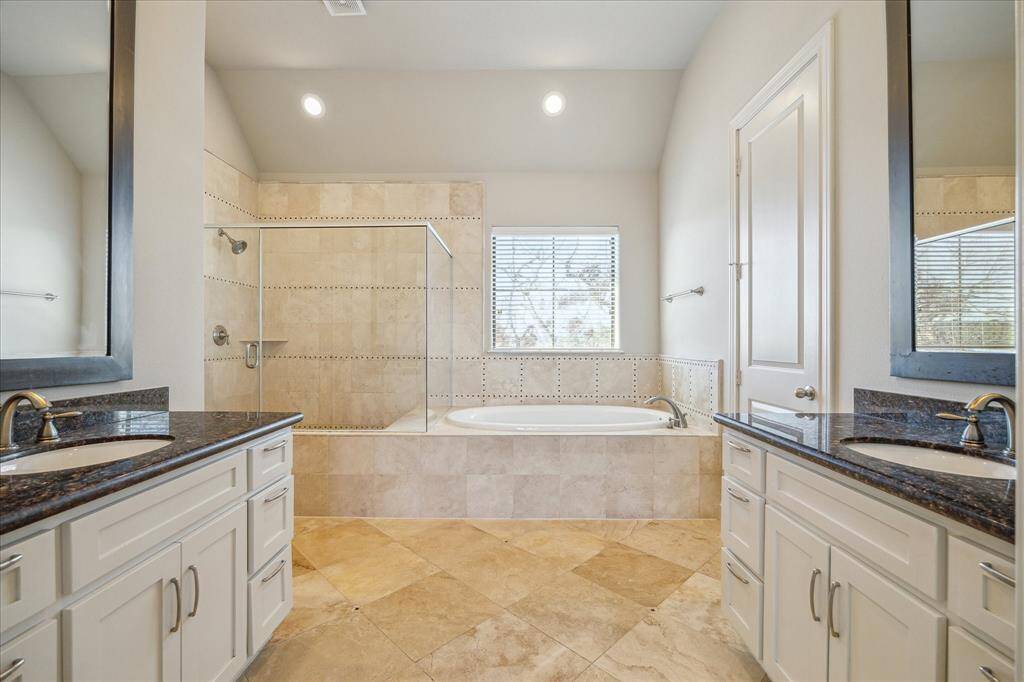 This spacious bathroom features dual sinks with elegant dark countertops, a large soaking tub, and a separate glass-enclosed shower. The neutral tile flooring and ample natural light create a relaxing atmosphere.