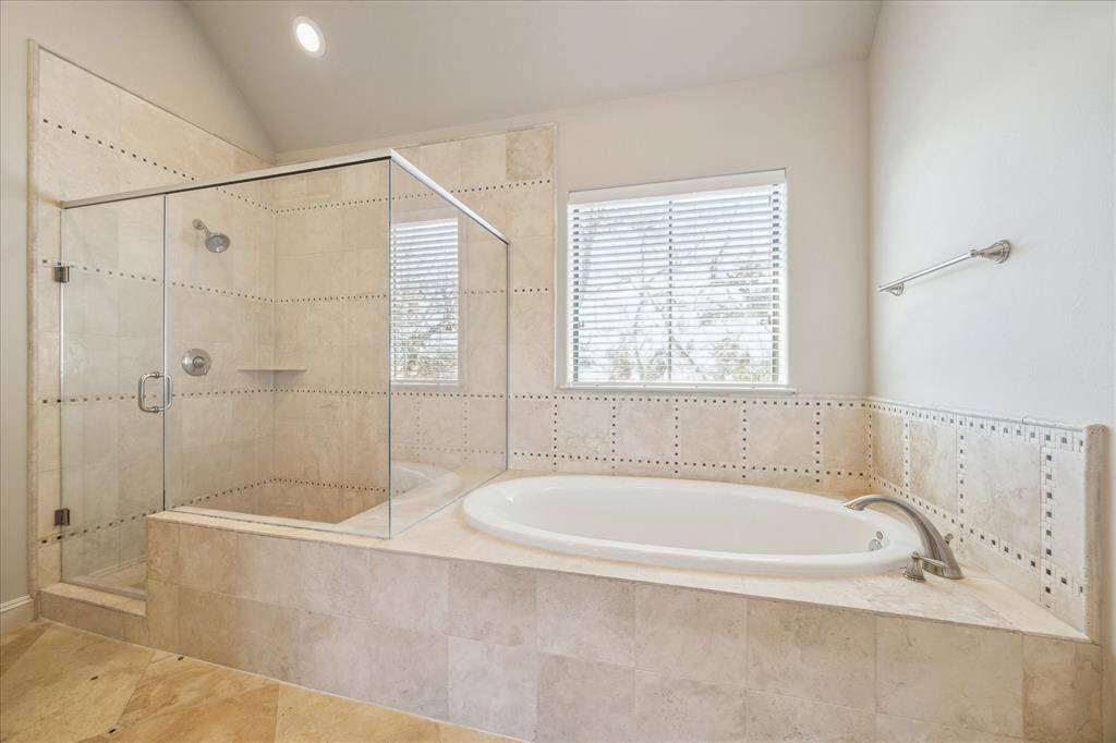 This bathroom features a luxurious design with a spacious glass-enclosed shower and a large soaking tub. The neutral tiles and natural light from the window create a serene atmosphere.