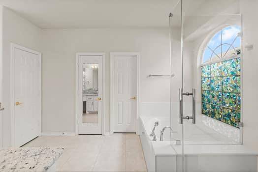 Spacious bathroom with a glass-enclosed shower and a large soaking tub beneath an arched window with decorative glass. The room features a neutral color palette, creating a bright and airy atmosphere.