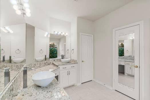 This modern bathroom features granite countertops, a vessel sink, ample mirror space, and bright lighting. The white cabinetry and neutral tones create a clean, spacious spa like feel.