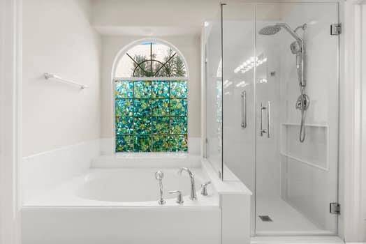 This bathroom features a luxurious design with a spacious bathtub and a modern glass-enclosed shower. A large, arched window with decorative glass allows natural light to fill the room, creating a bright and inviting atmosphere.
