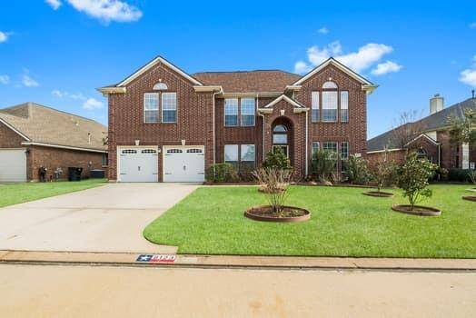 This is a spacious two-story brick house featuring a well-maintained front lawn with landscaped plants. It has a two-car garage and a welcoming entrance, perfect for a family home in a suburban neighborhood.