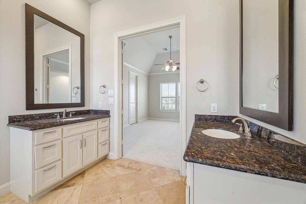 This photo showcases a spacious bathroom with dual sinks, granite countertops, and large mirrors. The room has a neutral color palette and connects to a bright bedroom area with carpeted flooring.