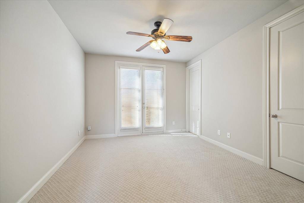 This photo showcases a bright, empty room with neutral walls and carpet, featuring a ceiling fan with lights. The room has double doors with blinds, allowing natural light to filter through, creating a cozy atmosphere.