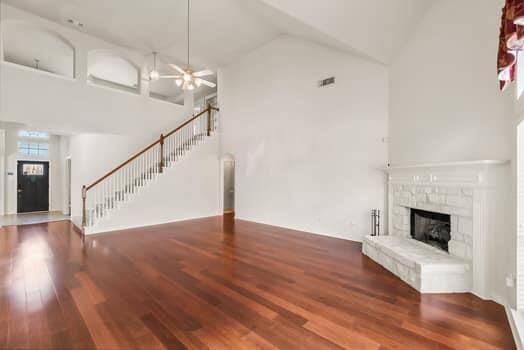 This spacious living area features beautiful hardwood floors, a high ceiling with a ceiling fan, and a white stone fireplace. The room includes an elegant staircase leading to an upper level, with ample natural light and a modern, open layout.