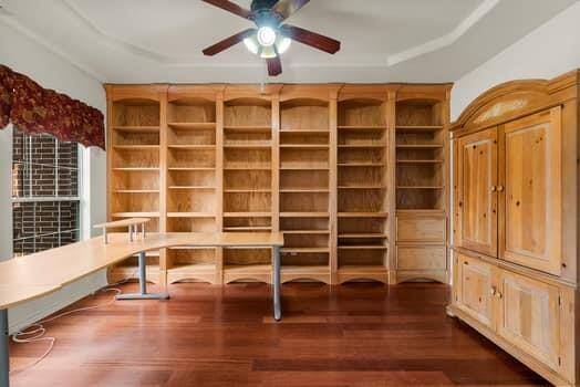 This room features warm hardwood flooring, built-in wooden bookshelves, and ample natural light from large windows. A ceiling fan provides added comfort, making it ideal for a home office or study.