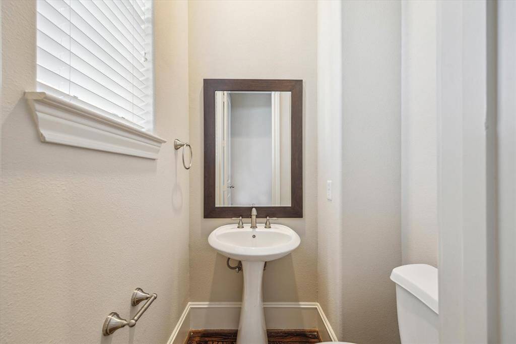 This photo shows a compact, well-lit powder room featuring a pedestal sink, a framed mirror, and a window with blinds for natural light on the second floor by living area. and kitchen.