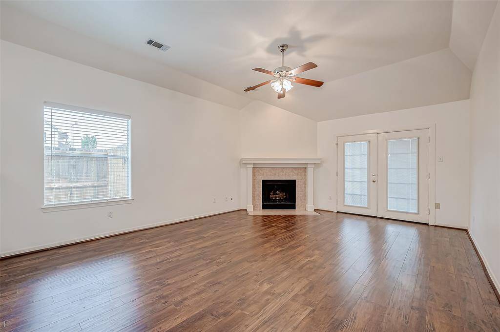 A large window allows sunlight to pour into the living room, highlighting the contrast between the light walls and the darker wood-like flooring. The corner fireplace serves as a cozy focal point, positioned beside elegant double doors that offer additional access.