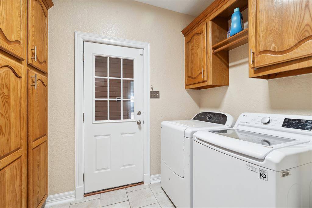 Compact laundry room with tiled flooring, featuring a washer and dryer. It includes wooden cabinets for storage and a door with a window for natural light.