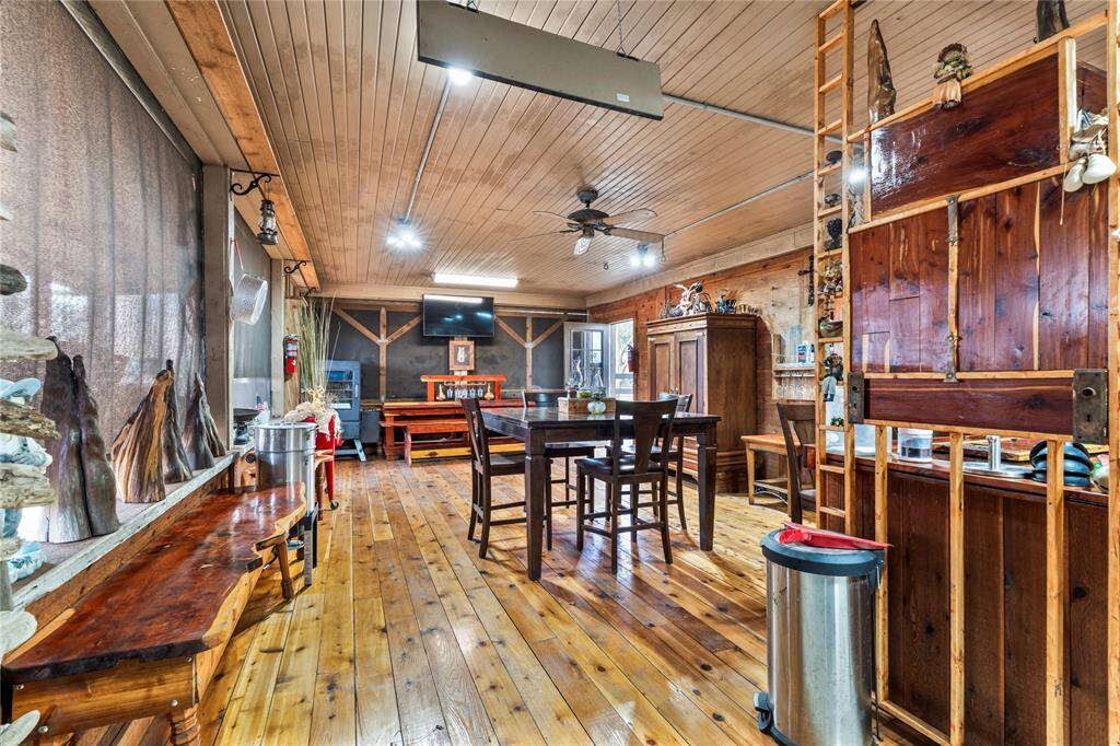 Rustic, cabin-like design with wooden floors, walls, and ceiling. It includes a dining area, a ceiling fan, and various wood furnishings. Natural light filters through a large window, creating a cozy, inviting atmosphere.