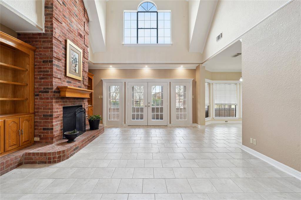 This spacious living area features high ceilings and large windows with lots of natural light. Beautiful, traditional brick fireplace, built-in wooden shelving, and French doors leading to an outdoor space. The neutral tile flooring enhances the room's bright and open feel.