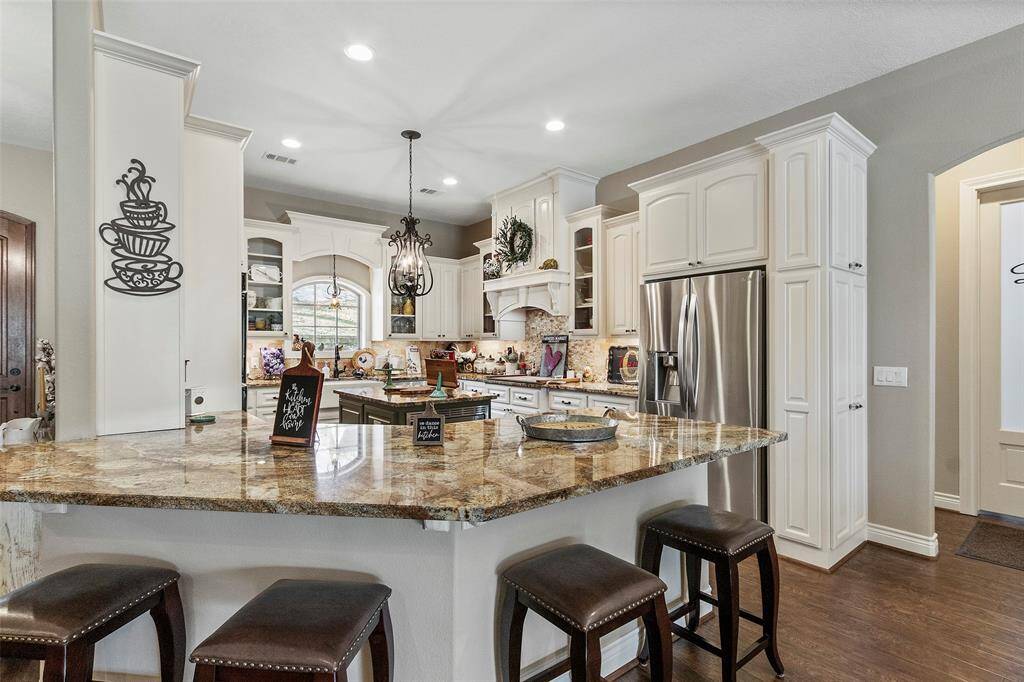 This kitchen features elegant white cabinetry, a large granite island, breakfast bar, and stainless steel appliances. It has a warm, inviting atmosphere with stylish lighting and decorative accents, perfect for both cooking and entertaining.
