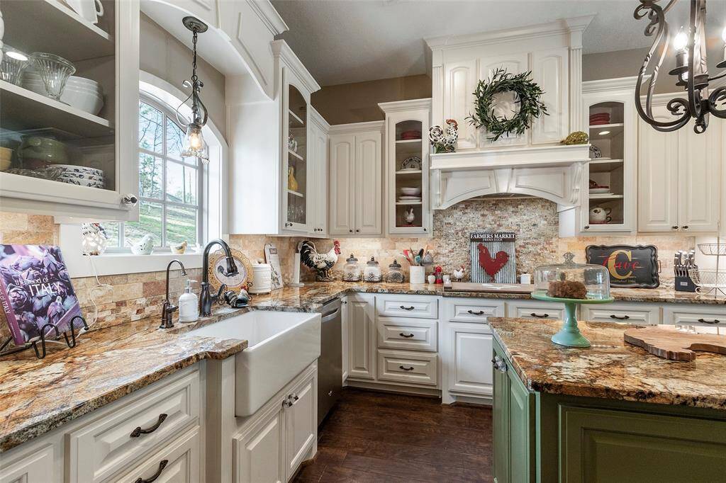 This charming kitchen features elegant white cabinetry, a farmhouse sink, and stunning granite countertops. A rustic stone backsplash complements the warm-toned granite, while glass fronts cabinets offers display space. The room is well-lit with a large window and a stylish chandelier, creating a cozy and inviting atmosphere.