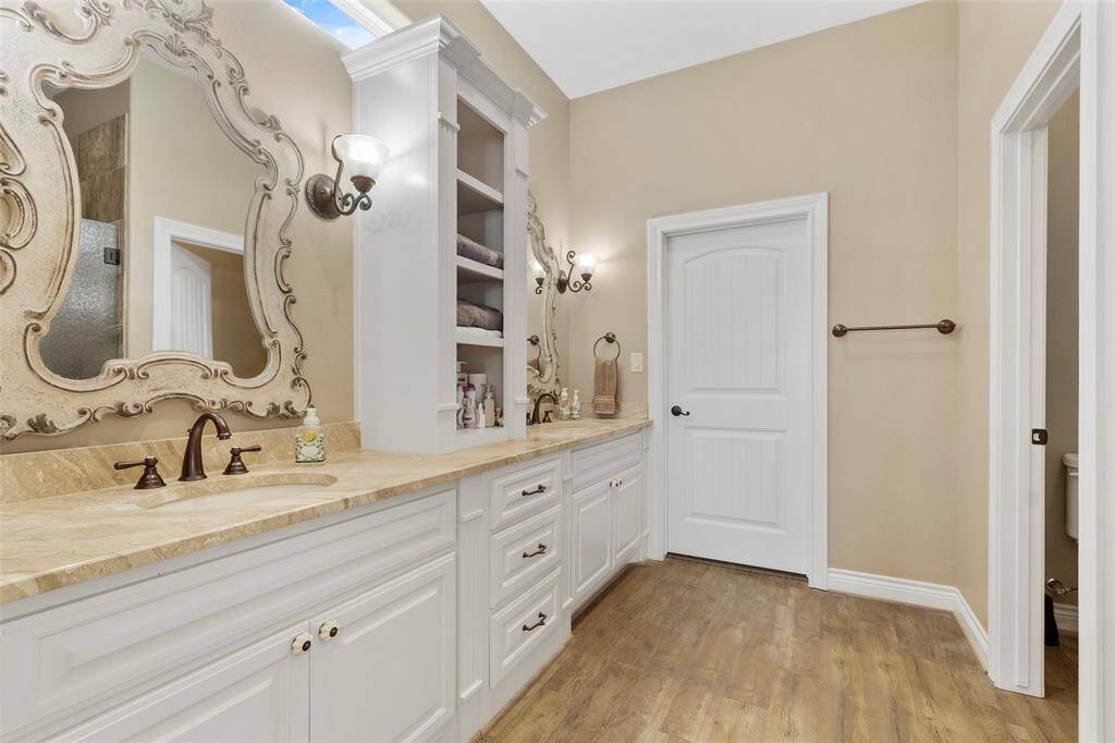 This elegant bathroom features dual sinks with darling ornate mirrors, a granite countertop, and ample cabinetry. The warm-toned luxury vinyl wood flooring complements the neutral wall color, creating a cozy and relaxing atmosphere.