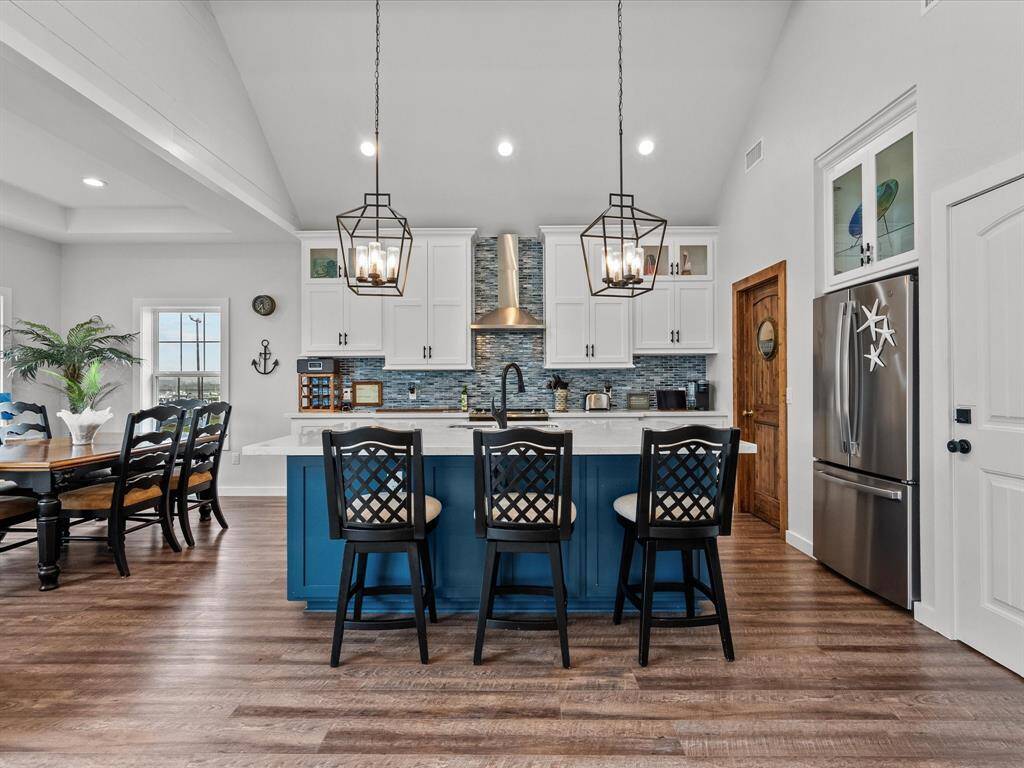 This photo showcases a modern kitchen with a spacious open layout. It features a large island with seating, stylish pendant lighting, and stainless steel appliances. The cabinetry is white with a blue tile backsplash, complementing the wooden floors. Adjacent is a cozy dining area with ample natural light.