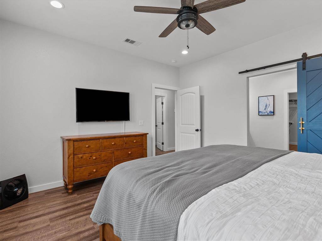 This bedroom features a cozy layout with a ceiling fan, a mounted TV, and a wooden dresser. It has laminate wood flooring, a white door leading to an adjacent room, and a stylish blue sliding barn door.