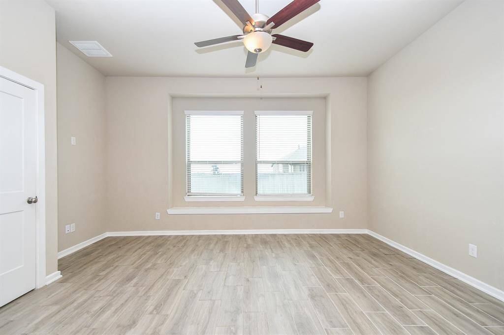This spacious master bedroom features light wood-look tile flooring, neutral walls, a large window with blinds, and a ceiling fan with lights, offering a bright and airy feel.