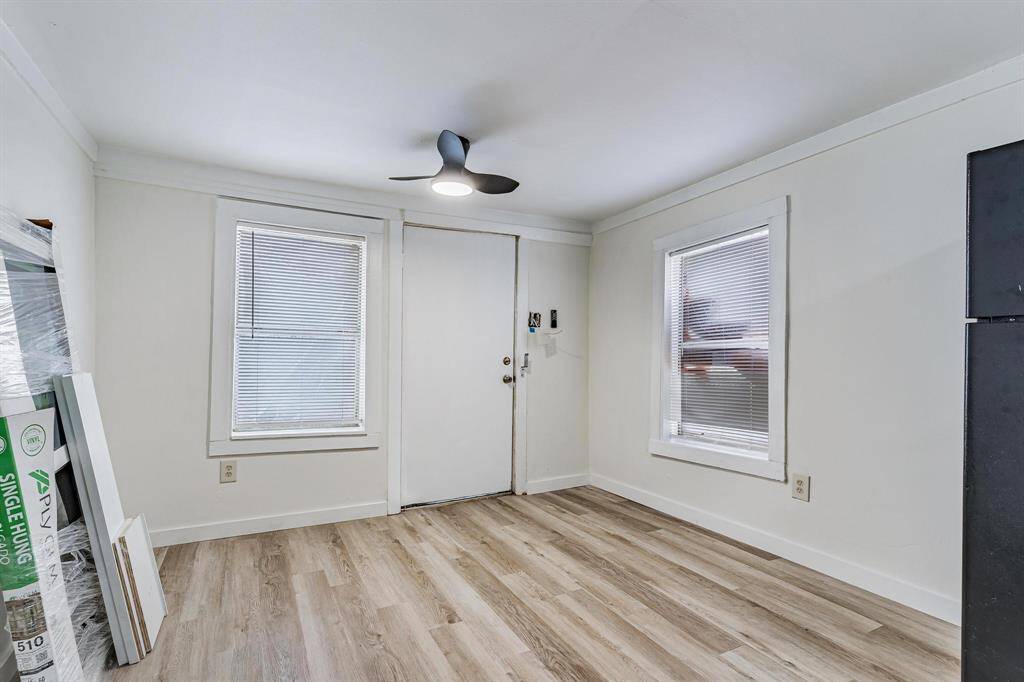 This is the view from the kitchen to the front door -  a comfortable, bright room with light wood flooring, white walls, and two windows with blinds. It features a modern ceiling fan and a simple door, creating a clean and minimalistic space.