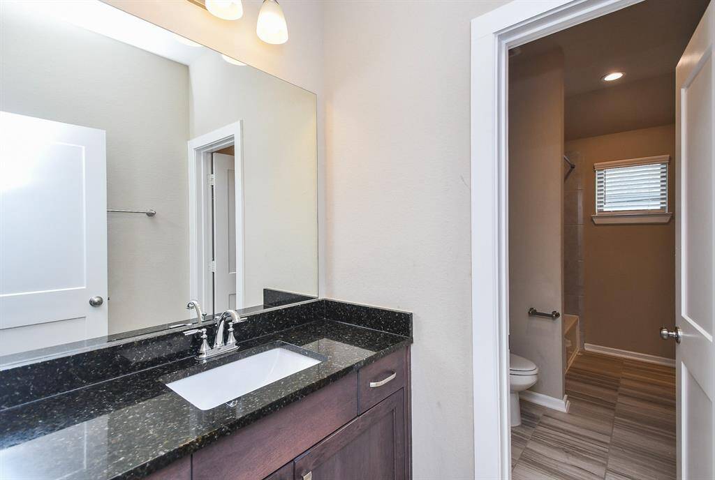 This third full attached bathroom upstairs features a sleek black granite countertop with a modern sink and faucet. It includes ample under-sink storage and is well-lit with overhead lighting. The adjoining area has a tiled floor, a small window, and a shower, creating a functional and stylish space.
