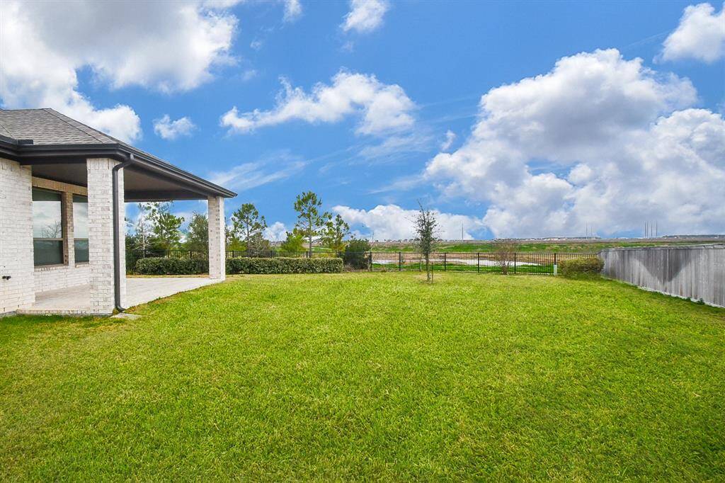 This photo showcases a spacious backyard with lush green grass, a covered patio area attached to a brick house, and a water view and trees under a bright, cloudy sky. Ideal for outdoor activities and relaxation. Yard is big enough for a swimming pool.