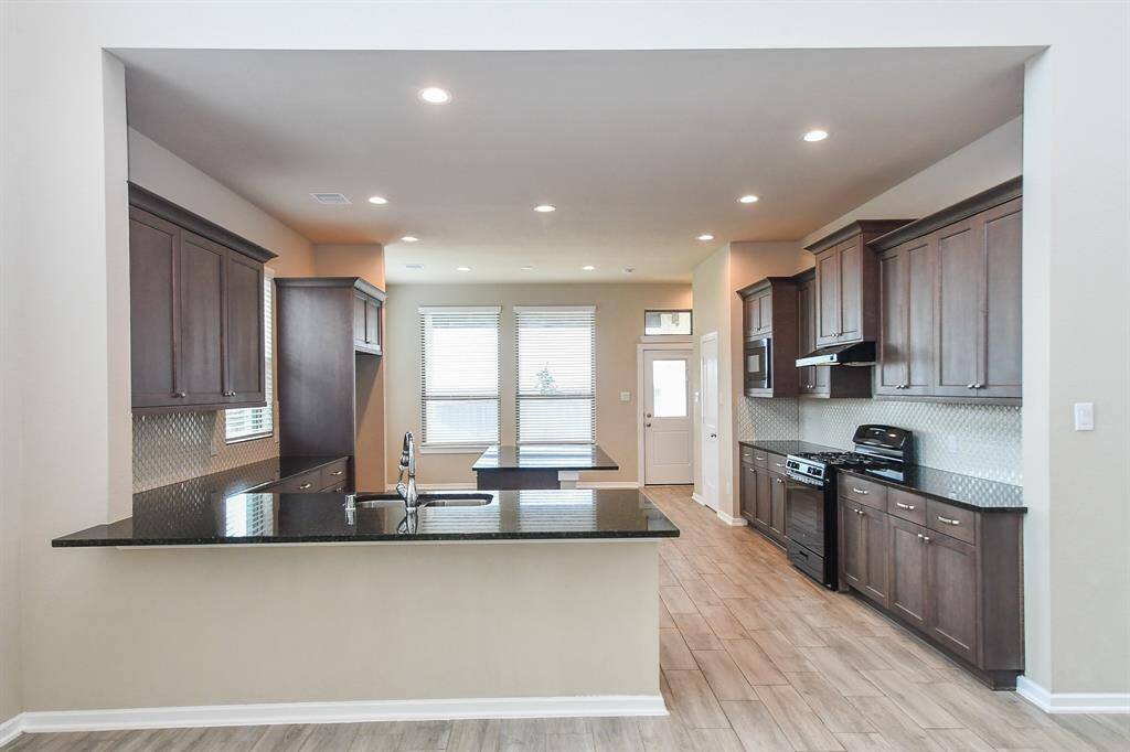 The kitchen features dark wood cabinetry, a sleek black countertop, and modern appliances. Natural light fills the space through large windows, complementing the neutral wall color and tiled floor.