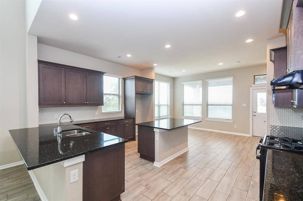 This kitchen features modern dark wood cabinets, granite countertops, and a central island with a sink. It has ample natural light from large windows and an open layout, perfect for cooking and entertaining.