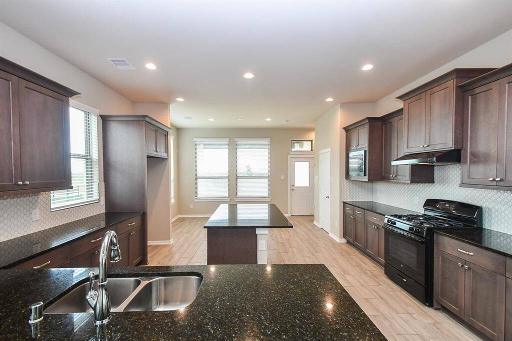 This spacious kitchen features elegant dark wood cabinets, sleek granite countertops, and modern appliances. It includes a central island for additional workspace and storage. The room is well-lit with recessed lighting and natural light from large windows, creating a bright and welcoming atmosphere.