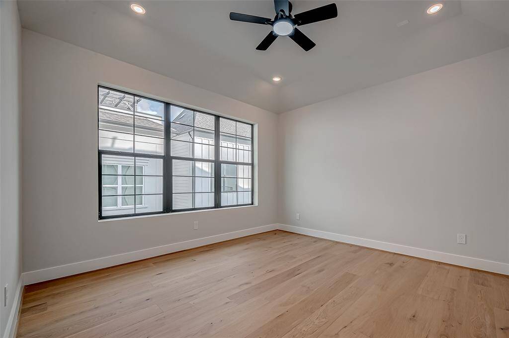 Bright room with large windows, light wood flooring, and neutral walls. Features a ceiling fan and recessed lighting. There's an open doorway leading to a hallway. Ideal for a bedroom or office space.